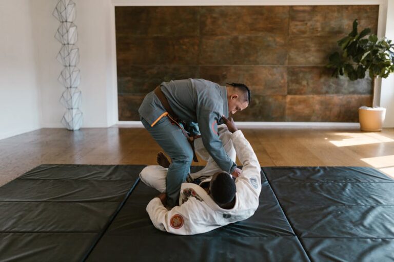 Two martial artists are practicing grappling techniques on black mats in a well-lit studio. One practitioner is in a grey gi and standing over the other in a white gi, who is lying on their back. The room has a wooden floor and a plant in the background.