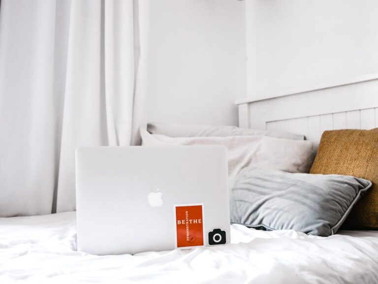 A white bed with white pillows and a mustard yellow cushion. On the bed is an open silver laptop with an Apple logo, a red card with "BE THE" text, and a small black digital camera. The setup is minimalist and cozy with white curtains in the background.