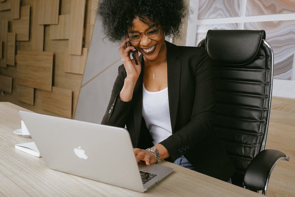 A person with curly hair and glasses is smiling while talking on the phone and using a laptop. They are wearing a black blazer over a white top and are seated at a wooden desk with a leather chair in an office setting.