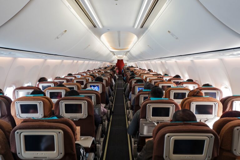 Interior view of an airplane cabin showing rows of passengers seated. Each seat has an individual screen on the backrest and passengers are engaged with various activities. The aisle runs down the center of the plane. Light from the windows illuminates the space.