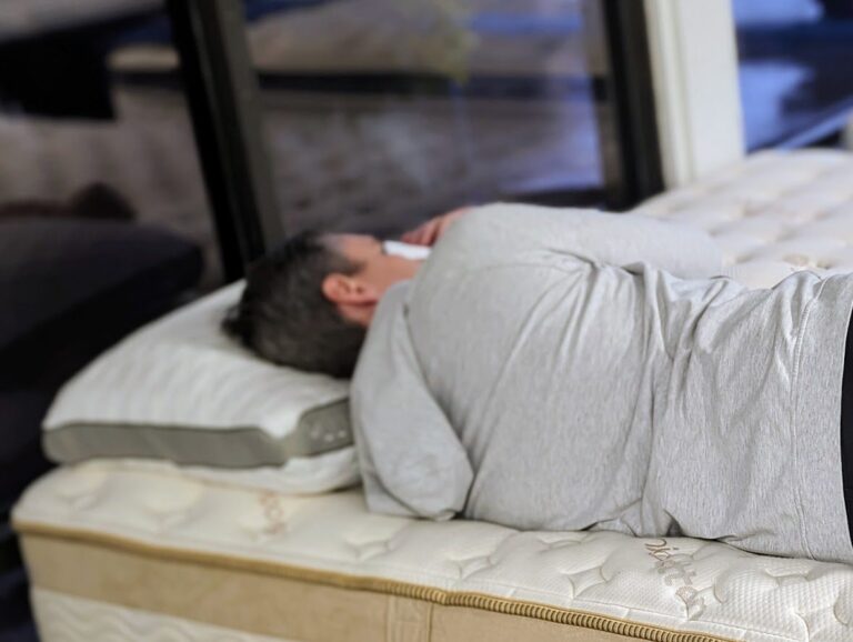 A person lying on their side on a mattress, wearing a gray long-sleeve shirt. The room has large windows in the background, and the mattress has a light-colored cover with a patterned design. The person rests their head on a pillow.