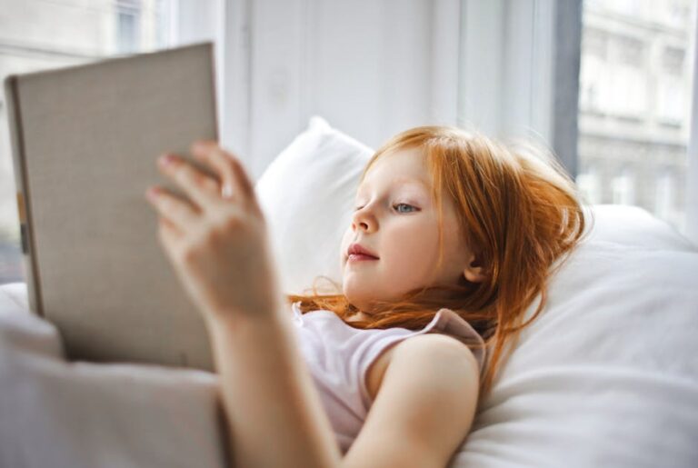 A young girl with long red hair is lying on a white bed, reading a book. She is propped up with white pillows and appears focused on the book she is holding. Natural light filters through a nearby window, illuminating the scene.