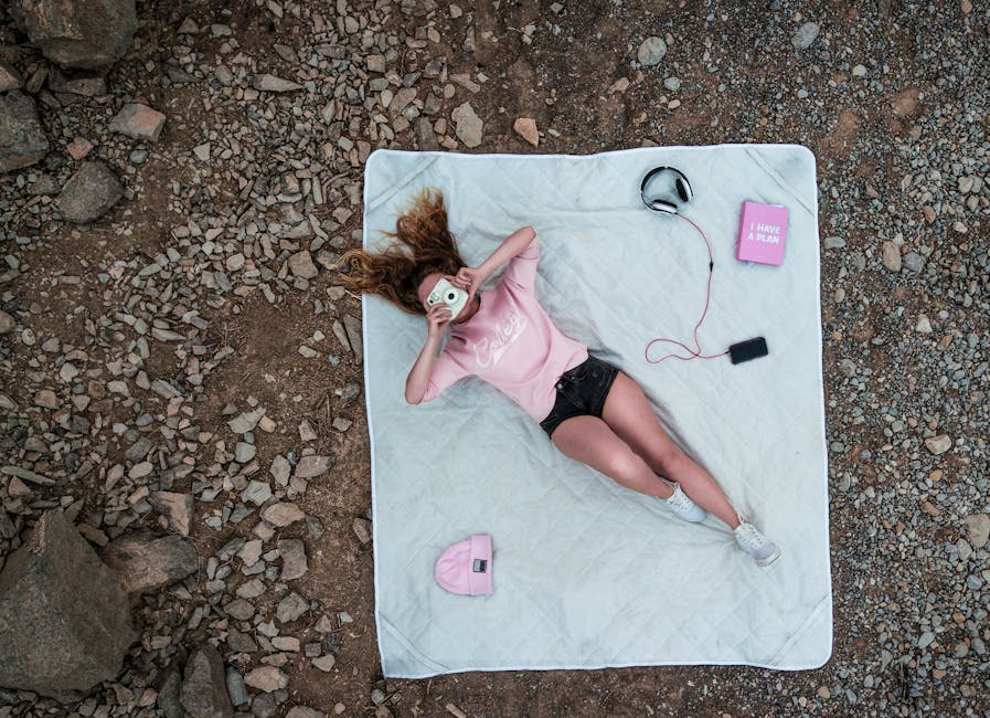 A person lies on a white blanket on a rocky ground, holding a camera to their face. They wear a pink shirt, black shorts, white sneakers, and a pink beanie is placed nearby. Headphones and a book titled "I have a plan" are also on the blanket.