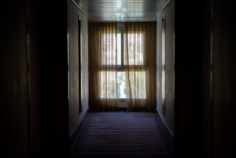 A dimly lit hallway with a striped carpet leads to a window covered by sheer, light brown curtains. Soft light filters through the curtains, casting a gentle glow in the otherwise dark corridor.