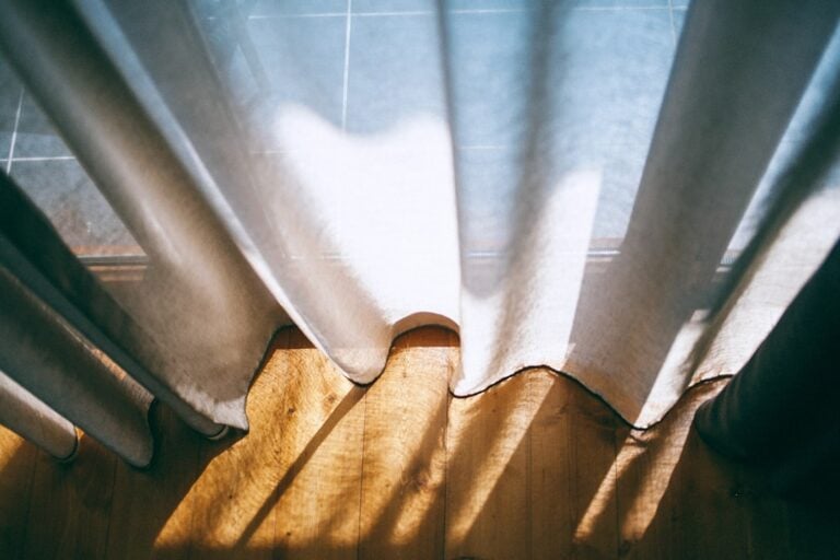 Light filters through sheer gray curtains, casting soft shadows on a wooden floor. The sunlit floorboards and the cool tile in the background create a contrast between warmth and cool tones in this tranquil scene.