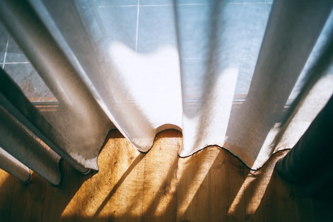 Light filters through sheer gray curtains, casting soft shadows on a wooden floor. The sunlit floorboards and the cool tile in the background create a contrast between warmth and cool tones in this tranquil scene.