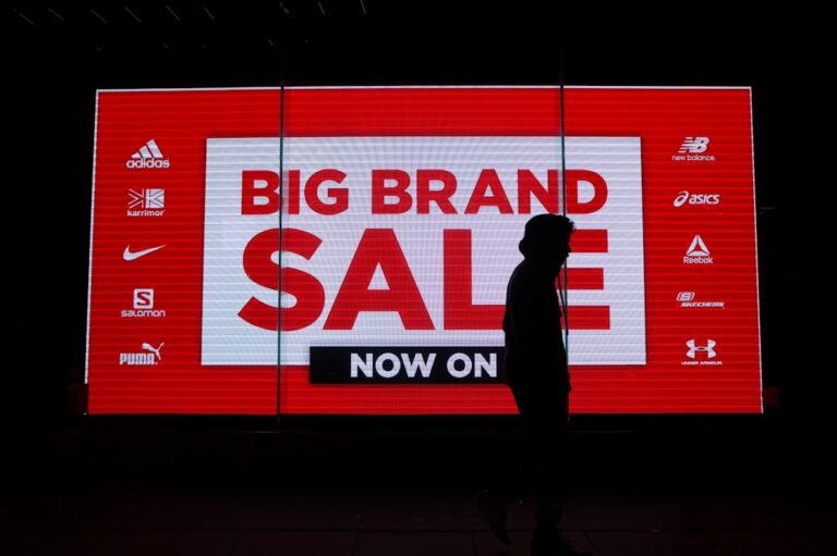 A large digital billboard displays "Big Brand Sale Now On" in bold white text on a red background, showcasing logos of various sports brands such as Adidas, New Balance, Puma, and Reebok. A silhouetted person walks in front of the sign.