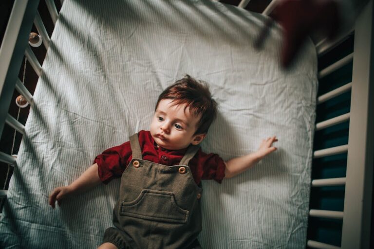 A baby with dark hair wearing a red shirt and grey overalls lies on their back in a crib with light-colored bedding, looking slightly to the side. The crib bars and shadows create a pattern across the scene.