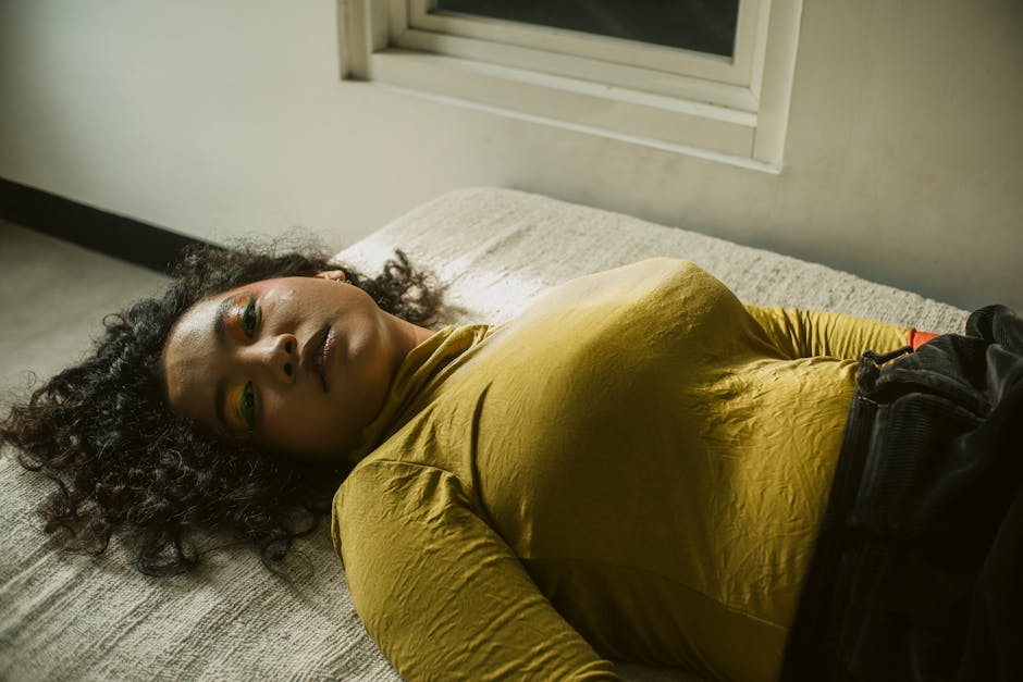 A woman with curly hair lies on a bed, wearing a mustard yellow top. She has colorful eyeshadow and a relaxed expression. A window is visible in the background, casting natural light into the room. The overall mood appears calm and contemplative.