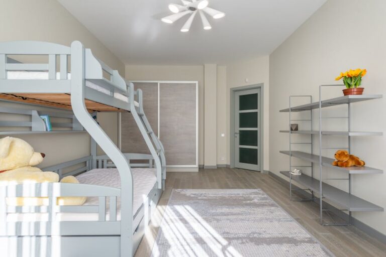 A tidy, modern room with light-colored walls features a bunk bed on the left, a plush teddy bear on the lower bed, a sleek shelf on the right adorned with potted flowers and a stuffed animal, a large wardrobe, and a stylish ceiling light. Intricate rug covers wooden floor.