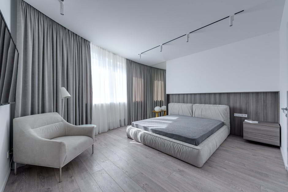 A minimalist bedroom with a large bed featuring a gray duvet, a modern beige armchair, a small wooden nightstand with a lamp, and floor-to-ceiling gray curtains covering large windows. The room has light wood flooring and overhead track lighting.