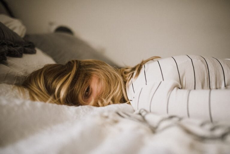 A person with long blonde hair is lying face-down on a bed, partially hidden by their hair. They are wearing a white shirt with black stripes. The room appears softly lit and the bedding is light-colored.