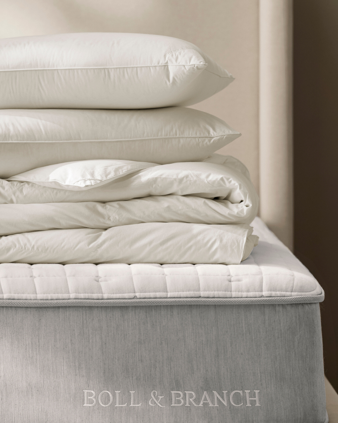 A neatly stacked arrangement of bedding, including three pillows and a folded comforter, is placed on the edge of a bed with a light grey mattress. The bedding and pillows are white, and the brand "Boll & Branch" is visible on the side of the mattress.