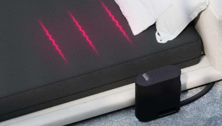 A black mattress with three red, wavy lines of light projected on its surface is placed on a beige bed frame. A black rectangular device with a connecting tube is positioned beside the mattress on the floor.