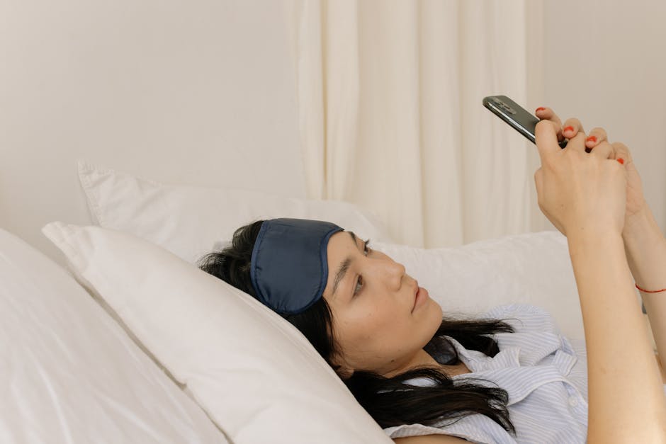 A woman lying in bed with white pillows, wearing a blue sleep mask on her forehead, looks at her smartphone with both hands. She appears to be relaxed and focused on the screen.