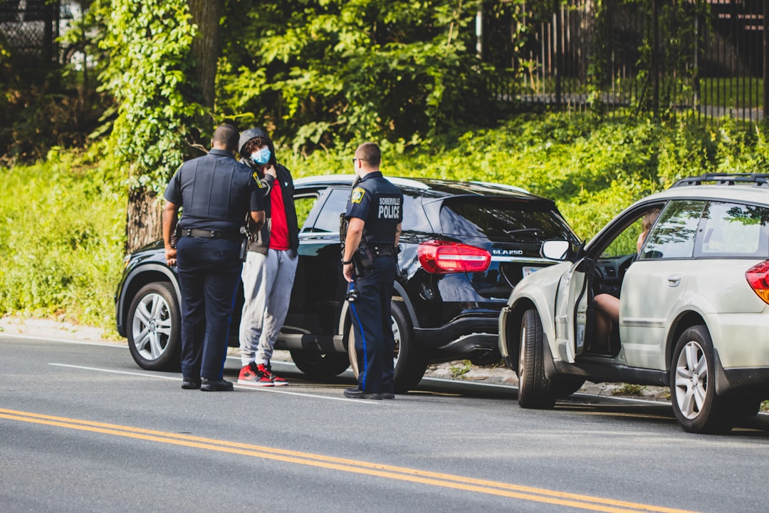 Two police officers are standing beside a black sedan that is pulled over on a street. They are engaging with a person in a red hoodie and gray pants, who is standing next to a black SUV. Another vehicle with an open door and a person inside is parked ahead. Trees are in the background.