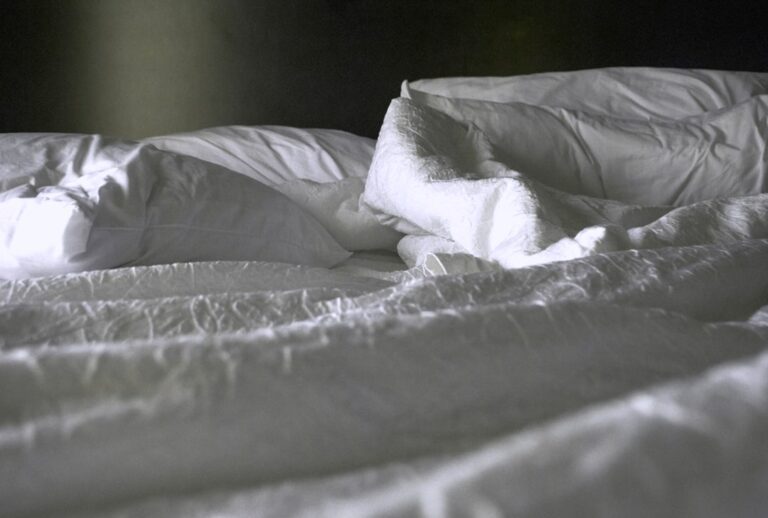 A rumpled bed with white sheets, pillows, and blankets. Light is softly illuminating part of the bedding, creating shadows and highlighting the texture of the fabric. The overall scene conveys a sense of calm and relaxation.