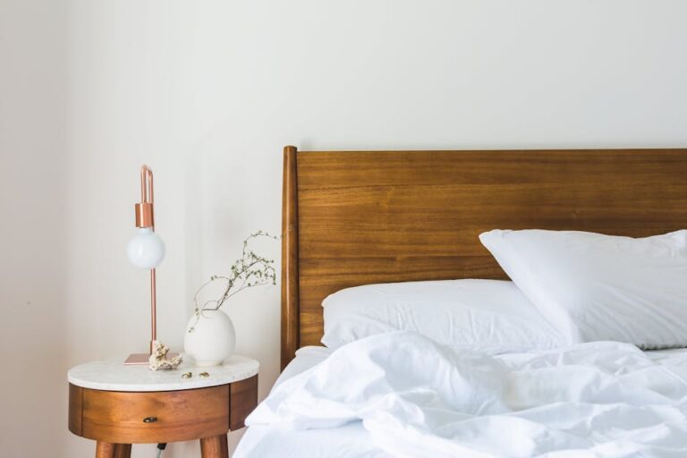 A minimalist bedroom scene featuring a wooden bed with white bed linens, a white pillow, and a round wooden side table. The table holds a white vase with dried branches and a modern copper lamp with a white bulb against a plain white wall background.