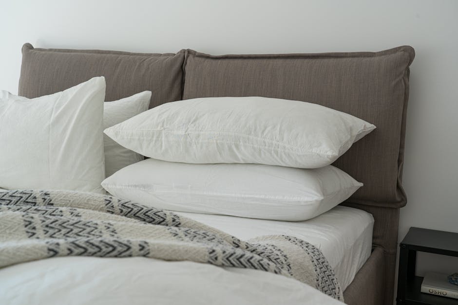 A neatly made bed with a gray headboard, two white pillows stacked on top of each other, and a patterned blanket. The bed is against a white wall, and a small black nightstand is partially visible on the right side.