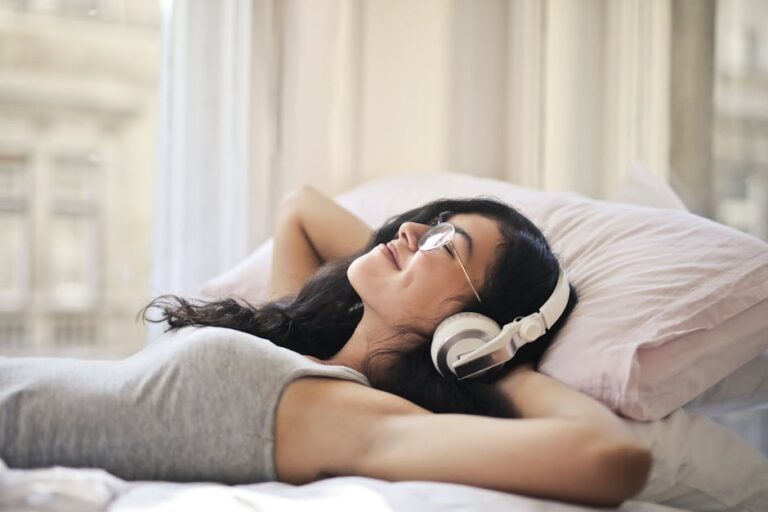 A person with long dark hair, wearing glasses and headphones, is lying on a bed with a content smile. They have their hands behind their head and are wearing a gray tank top. The room is lit by natural light from a nearby window.