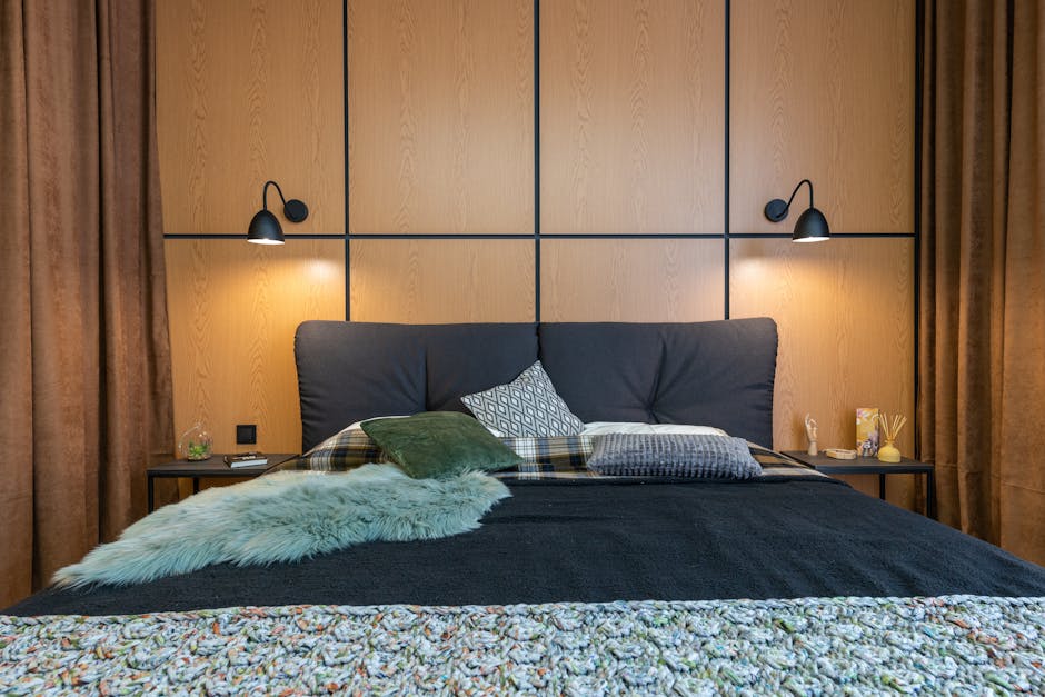 A cozy bedroom with a double bed featuring a mix of pillows and throws in green, white, and plaid patterns. Wall-mounted lamps with black shades illuminate the wooden headboard. Brown curtains frame the bed, creating a warm and inviting atmosphere.