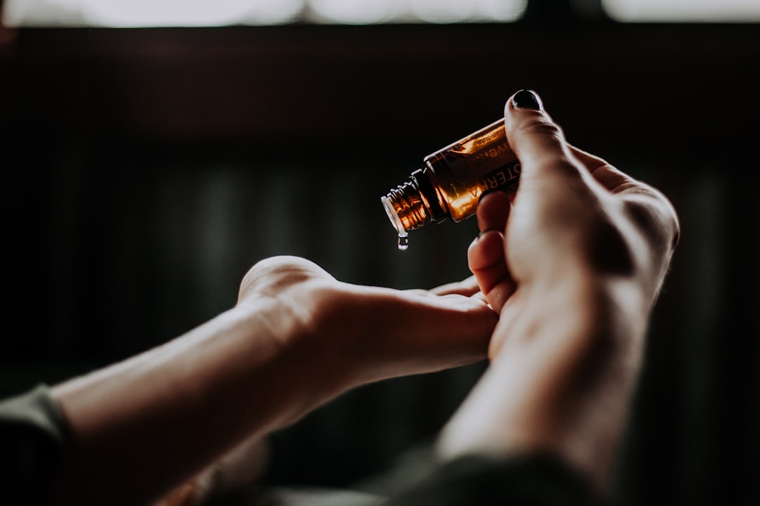 A close-up of hands holding a small brown bottle, tilted to pour oil or liquid into the palm of the other hand. The background is dark and blurred, emphasizing the focus on the hands and the bottle.