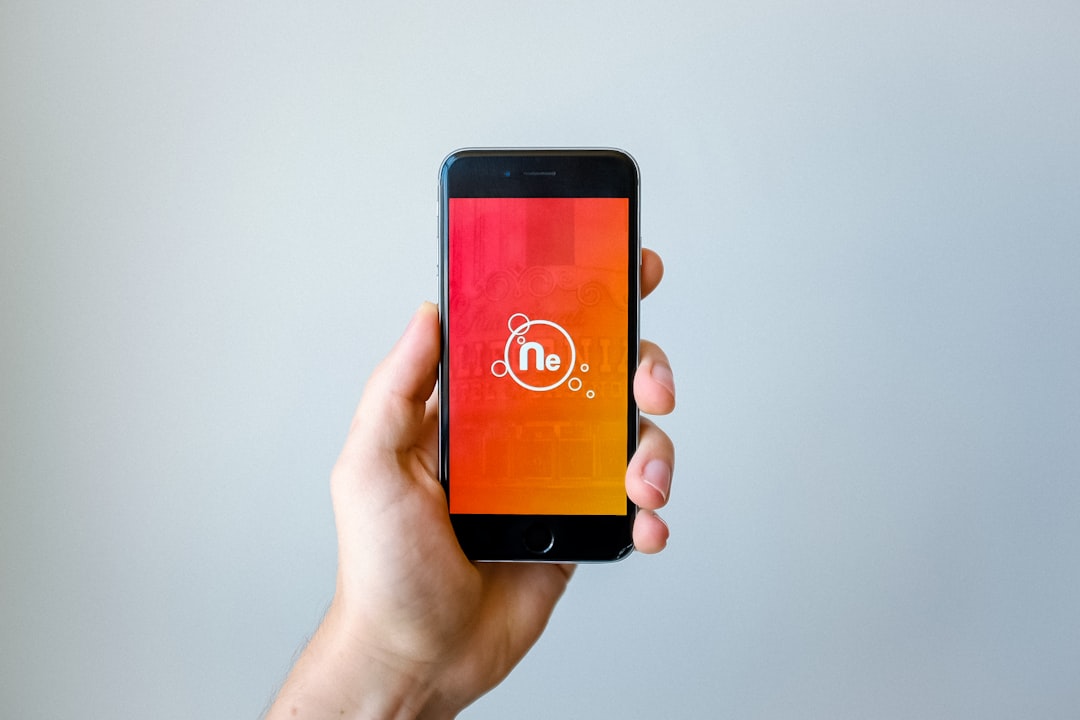 A hand is holding a smartphone with a screen displaying a red and orange gradient background. An "Ne" logo, encircled and decorated with small bubbles, is centered on the screen. The background is plain and light-colored.