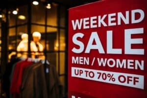 A red sign in a clothing store reads "Weekend Sale Men/Women Up to 70% Off." In the background, mannequins display clothing items, and racks of clothes are visible under ambient store lighting.