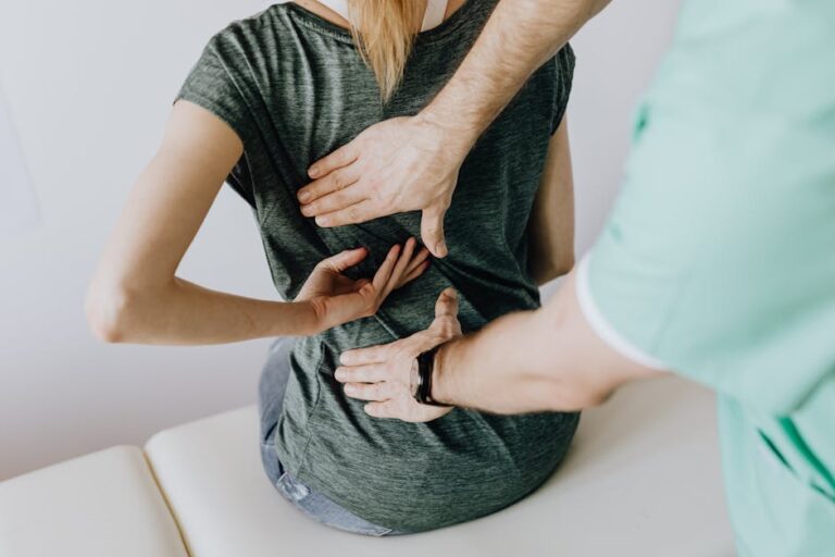 A person wearing a gray shirt is sitting on an examination table while a healthcare professional in a green outfit examines their back by placing hands on their spine and lower back, providing a possible chiropractic or osteopathic treatment.