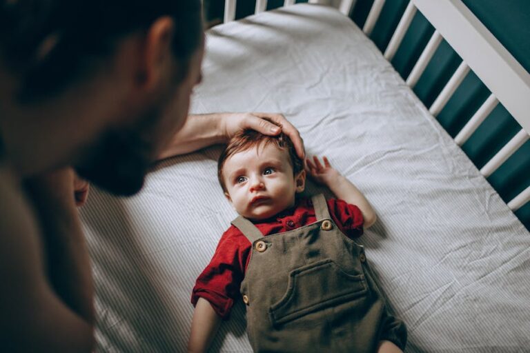 A baby wearing a red shirt and brown overalls lies in a white crib. An adult with a beard gently touches the baby's head while looking at them. The baby's eyes are wide open, looking up at the adult. The background is minimal, focusing on the crib and bedding.