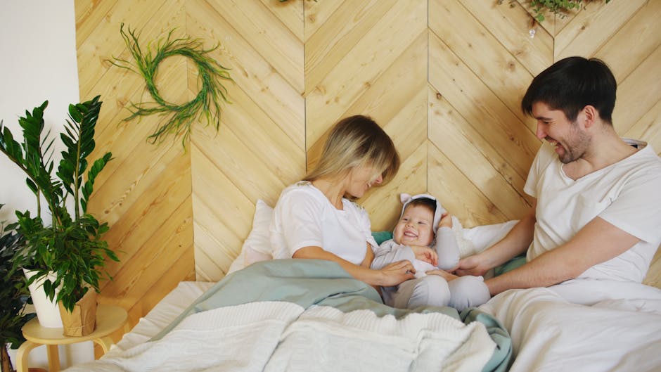 A family of three sits on a bed with a wooden headboard. The mother, on the left, gently touches their baby, who is dressed in white and sitting between the parents. The father, on the right, smiles and holds the baby’s legs. Green plants decorate the space.