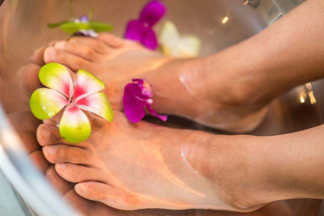 Close-up of two feet soaking in a water basin with colorful flower petals floating around. The water appears clear, and the flowers include vibrant purple and green petals, contributing to a relaxing, spa-like atmosphere.