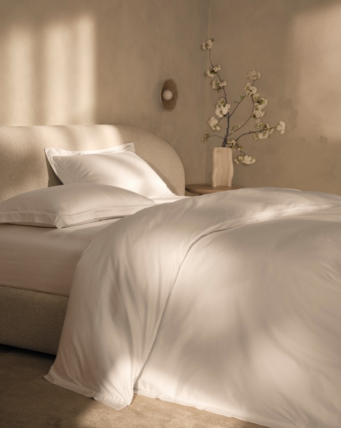 A cozy, minimalistic bedroom featuring a neatly made bed with white linens and pillows. Soft sunlight spills through the room, illuminating a nightstand with a minimalist vase holding a branch of white flowers. The overall ambiance is serene and inviting.