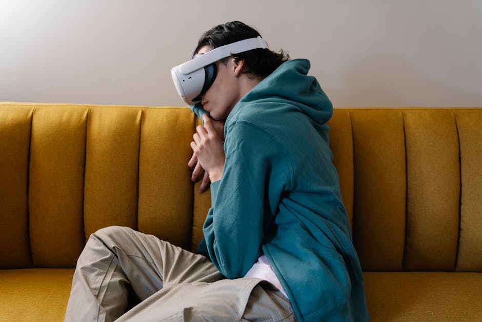 A person wearing a teal hoodie and khaki pants is seated on a mustard yellow couch. They are wearing a white virtual reality headset and appear to be deeply engaged in the experience, leaning forward with their hands clasped near their face.