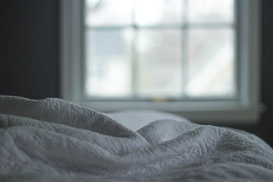 A cozy bed with white, crumpled covers in the foreground, positioned beside a window that is slightly out of focus. The soft light coming through the window illuminates the room, creating a peaceful and relaxing atmosphere.