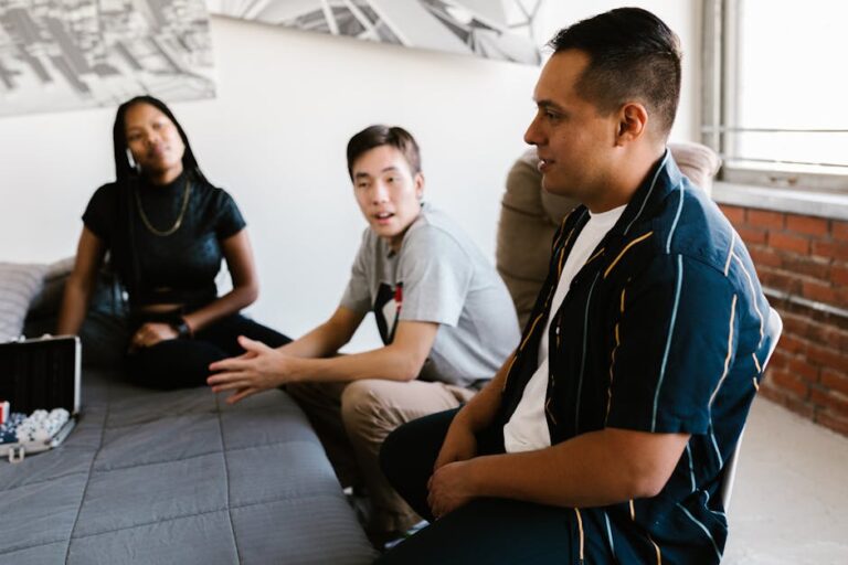 Three friends are gathered in a living room. One is seated on a bed, another on a chair, and the third is standing. They appear to be engaged in a conversation, with a poker set on the bed in front of them. The room has minimalistic décor with exposed brick walls.