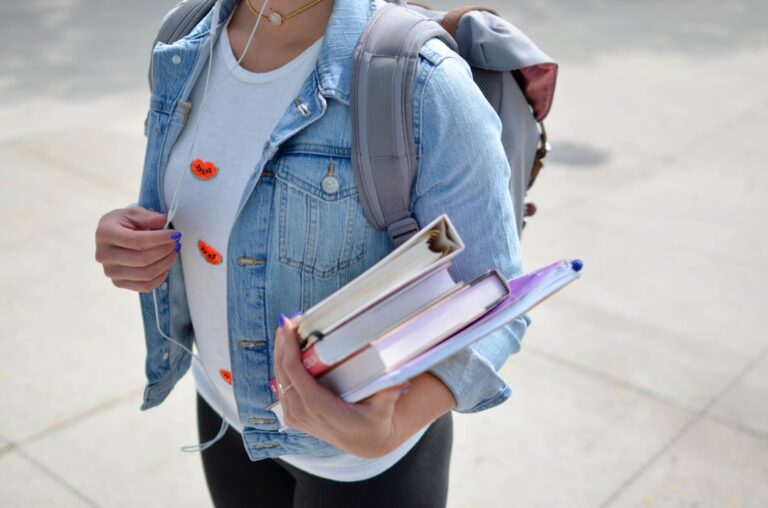 A person wearing a light denim jacket and a gray backpack holds several books while standing on a paved surface. Earphones are visible. The person's face is not shown.