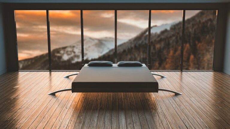 A minimalistic bedroom with a low-profile bed featuring two pillows. The room has floor-to-ceiling windows showcasing a scenic mountain landscape during sunset. The hardwood floor complements the modern decor, creating a serene and peaceful atmosphere.