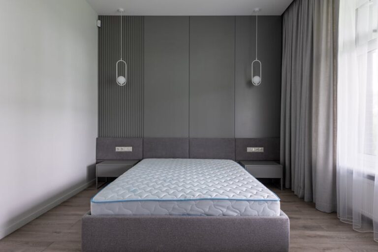 Minimalist bedroom with a grey upholstered bed frame and a mattress. The wall behind the bed features vertical paneling and two pendant lights. The room has grey curtains, two side tables, and ample natural light from a window on the right.