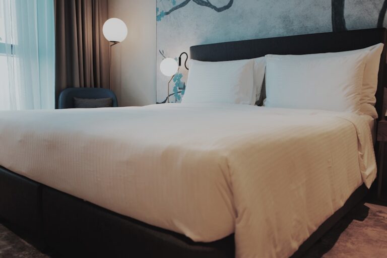 A modern hotel room with a large, neatly made bed featuring white linens and pillows. The room is softly lit by two spherical lamps, one on each side of the bed. A window with sheer curtains allows natural light to filter in, enhancing the room's cozy ambiance.