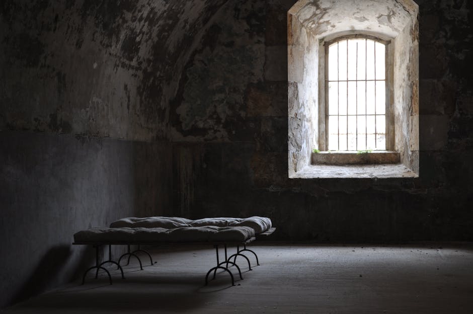 A dimly lit, rustic room with a rough, arched stone ceiling. There are two metal-framed single beds with thin mattresses on the left. A small barred window on the right allows sparse natural light to seep in, casting a faint glow on the aged, textured walls.