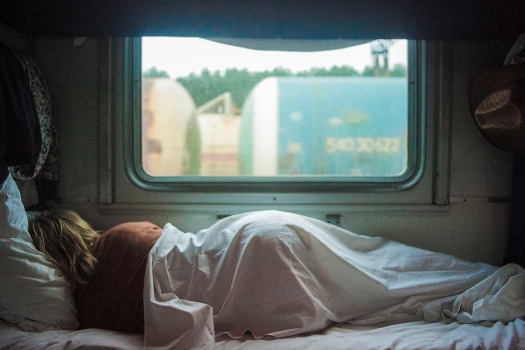 A person lies curled up under a blanket on a bed near a window in a train compartment. The view outside shows passing freight cars and greenery in the distance. An atmosphere of travel and relaxation is evident.