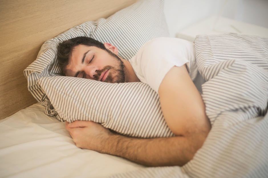 A man with short dark hair and a beard is sleeping in a bed. He is lying on his side, clutching a striped pillow under his head. The bedding, including the pillowcase and blanket, is also striped with a light color. The headboard is made of light-colored wood.