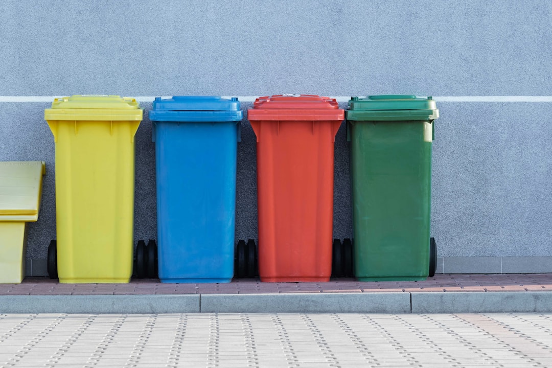 Four outdoor wheelie bins are lined up against a gray wall on a paved surface. The bins are color-coded in yellow, blue, red, and green, possibly for different types of waste recycling. A small portion of another yellow bin is visible on the far left.