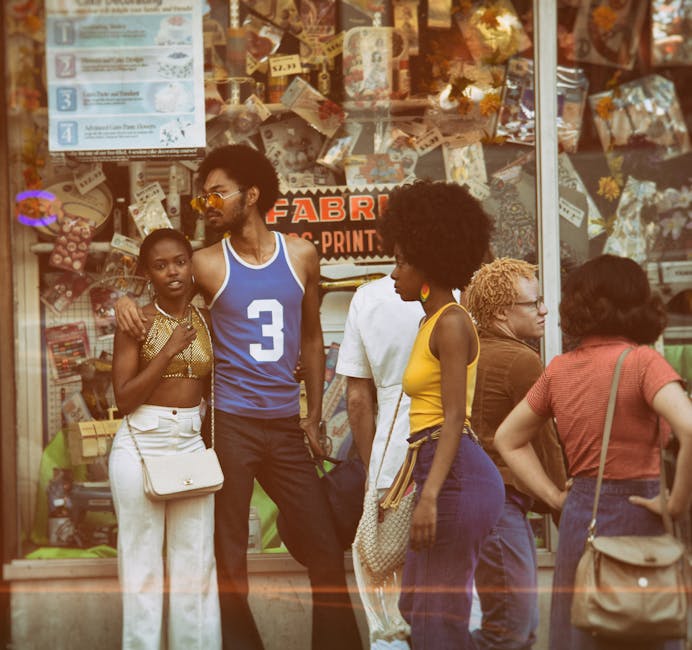 A group of people, dressed in vibrant 1970s fashion, stand and chat outside a fabric store with a busy, colorful display window. The store's sign reads "Fabrics" and people are engaged in conversation, capturing a moment of social interaction on the street.