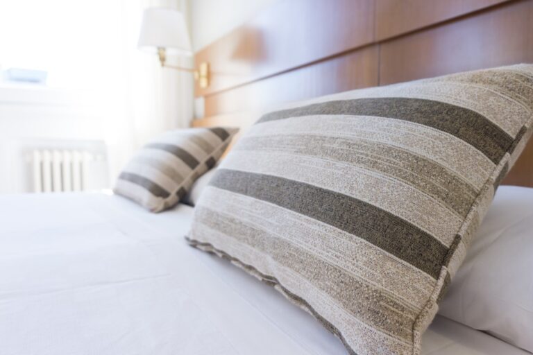 Close-up view of a neatly made bed with two striped pillows. The pillows have a light beige and dark brown horizontal stripe pattern. The headboard is wooden, and a lamp is mounted on the wall in the background. Sunlight filters through a nearby window.