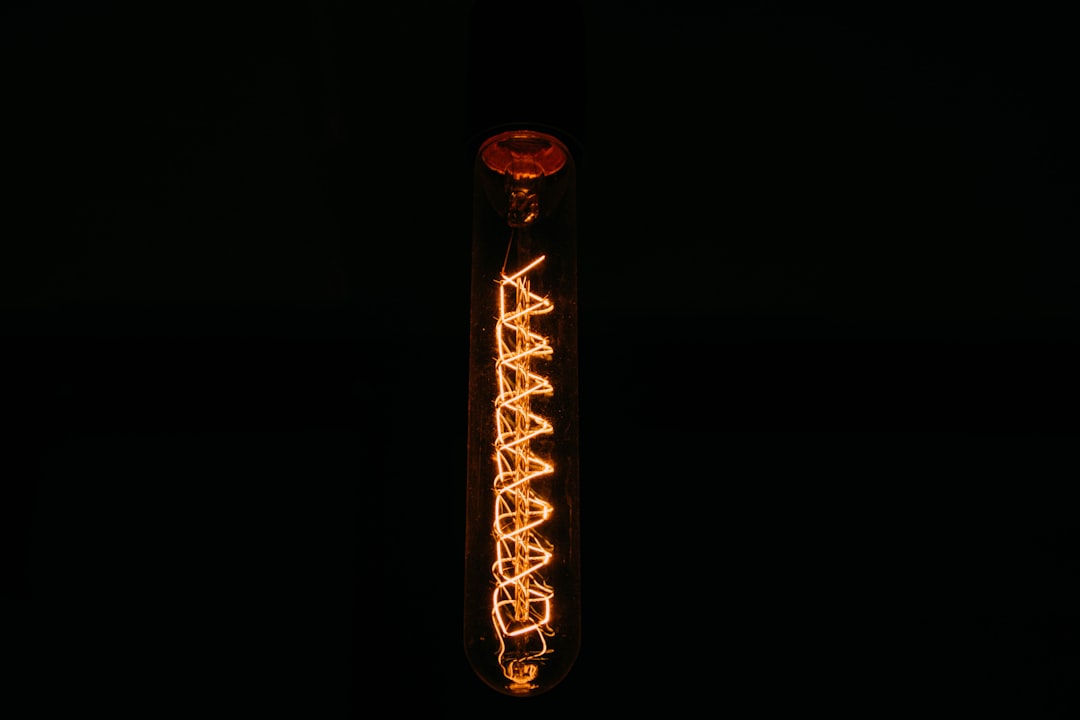 A single vertical light bulb against a dark background, with visible glowing filaments forming a zigzag pattern inside the bulb, casting a warm, orange light.