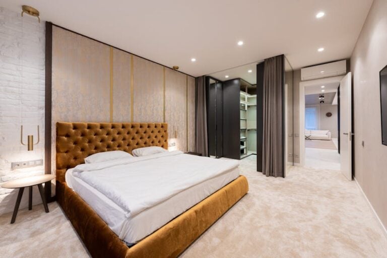 A modern bedroom with a large bed featuring a tufted brown headboard, white bedding, and two wooden nightstands with lamps. The room has recessed lighting, beige carpet, a built-in open closet with shelves, and a doorway leading to another room.