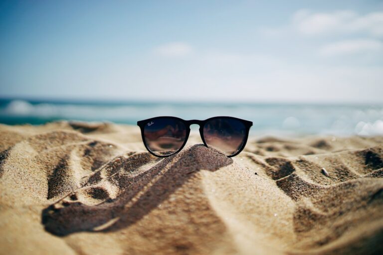 Sunglasses with dark lenses rest on a mound of golden sand on a sunny beach. The background features a clear blue sky and a distant, blurred ocean horizon, creating a relaxed and sunny coastal scene.