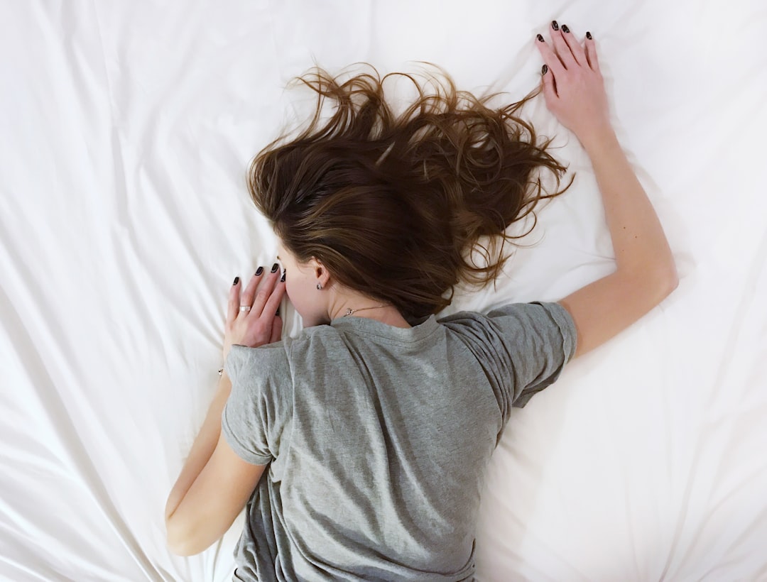 A person with long brown hair lies face down on a white bed. They are wearing a gray t-shirt, and their arms are spread out, with their hands by their head. Their nails are painted black. The scene conveys a sense of relaxation or exhaustion.